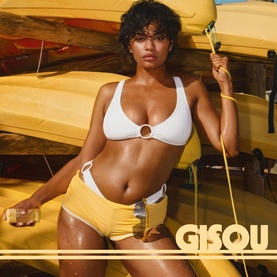 Gisou Body Mist held by model in white bikini top and yellow bottoms, posed outdoors near yellow kayaks