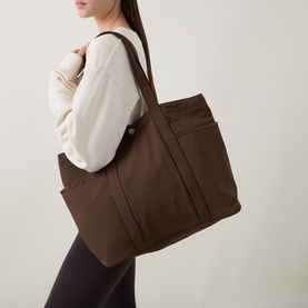 Model carrying Lululemon Daily Multi-Pocket Tote Bag in Walnut Crunch, worn over shoulder, styled with cream top and dark leggings.
