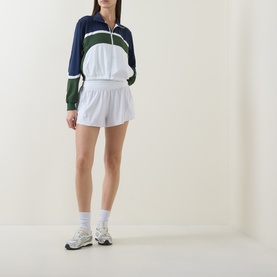Model wearing Lululemon Court Champ UV-Protective Tennis Jacket in White/Ivy Grove/Club Blue, styled with white tennis shorts and trainers.