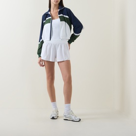 Model wearing Lululemon white shorts, styled with a navy and green colour-block zip-up jacket, white tank and chunky trainers.
