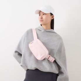Model with Lululemon Everywhere Belt Bag in Pink Pearl, rose gold hardware, worn crossbody over grey hoodie and cap