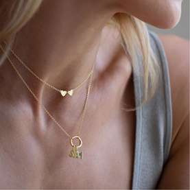 Model with Juvi gold layered necklaces featuring a small heart pendant and a charm holder with letter pendants, styled with grey vest top