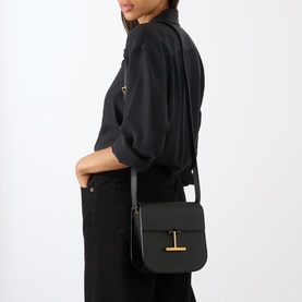 Model carrying Tom Ford Tara Mini Crossbody Bag in black grained calfskin with gold-tone T-bar hardware, worn crossbody over black outfit.