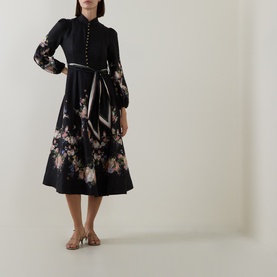 Model wearing Zimmermann Black Bouquet linen dress with floral hem, button front, balloon sleeves, and tie waist, styled with gold sandals.