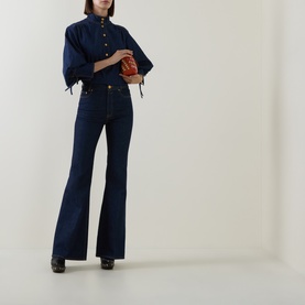 Model wearing Zimmermann Harbour Blue flare jeans, styled with matching denim shirt and black heeled boots, holding small tan bag.