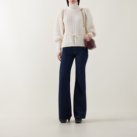 Model wearing Zimmermann Bubble Textured Turtleneck Sweater in cream with puff shoulders and front tie, styled with dark flare jeans and small purple