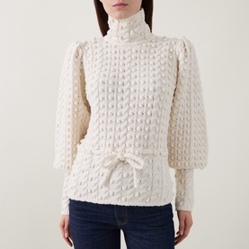 Model wearing Zimmermann Bubble Textured Turtleneck Sweater in cream with puff shoulders, blouson sleeves, and front tie, styled with blue jeans.