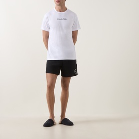 Model wearing Calvin Klein white cotton T-shirt with blue logo, styled with black shorts and dark slippers.
