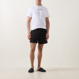 Model wearing Calvin Klein white cotton T-shirt with blue logo, styled with black drawstring shorts and navy slippers.