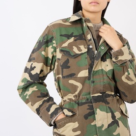 Model wearing green and brown camouflage utility jacket with chest pockets and snap fastening, styled with grey crew neck top