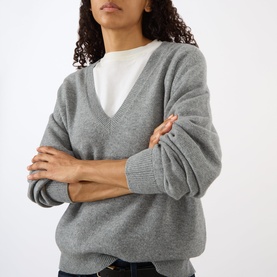 Model wearing Nili Lotan pebble grey heather v-neck knit sweater, styled over a white layering top, arms crossed.