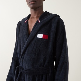 Model wearing Tommy Hilfiger Flag Logo Belted Robe in navy, showing hooded design, red and white striped trim, and embroidered flag logo pocket