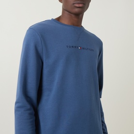 Model wearing Tommy Hilfiger blue crew neck sweatshirt with embroidered chest logo, cropped view showing torso and neckline.