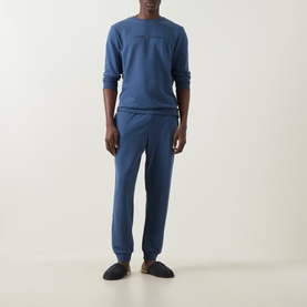 Model wearing blue crew neck sweatshirt and matching joggers, styled with dark slip-on slippers