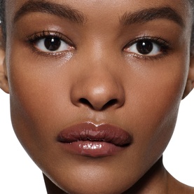Model wearing Victoria Beckham Posh Gloss in Bikini, a deep brown-nude lip gloss with a glossy finish, shown in a close-up beauty shot.