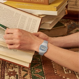 Model with Casio silver digital watch with mesh bracelet strap, worn on wrist against a backdrop of stacked books.