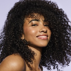 Model with voluminous, defined curly hair styled with Fenty Hair Dry Shampoo, smiling against a light background