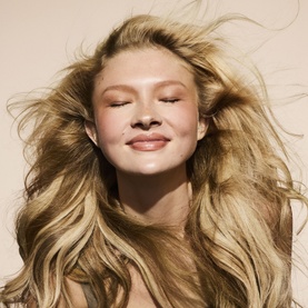 Model with voluminous, windswept blonde hair, eyes closed, smiling against a warm neutral background, showcasing Fenty Hair Dry Shampoo results