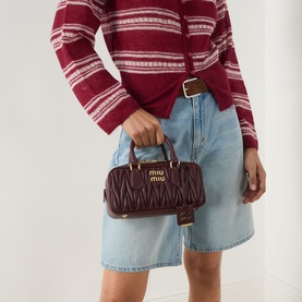 Model carrying Miu Miu Arcadie Matelass&eacute; Small Top Handle Bag in bordeaux with gold-tone hardware, styled with a striped knit and denim shorts.