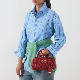 Model carrying Miu Miu Arcadie Matelass&eacute; Top Handle Bag in Rosso with gold-tone hardware, styled with a blue striped shirt and green scarf.