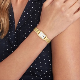 Model with Tommy Hilfiger gold-tone watch, rectangular white dial on a gold bracelet strap, styled with a navy polka-dot top.