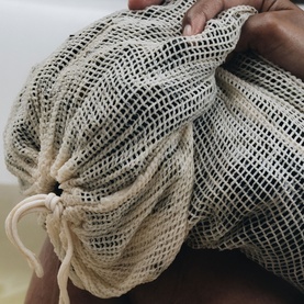 Voya mesh drawstring bag in natural ecru, held in hands, showing open-weave texture and drawstring closure detail.