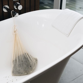 Voya mesh bath bag filled with seaweed, hanging from a freestanding white bathtub in a spa-style bathroom