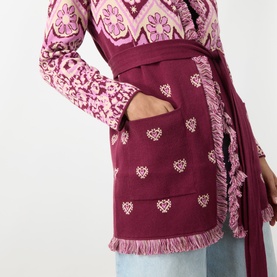 Model wearing burgundy embroidered wrap cardigan with heart motifs, fringe trim, and belt, styled over floral print top and blue trousers.
