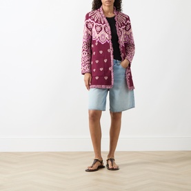 Model wearing pink and burgundy patterned jacquard cardigan coat with fringe hem, styled with denim shorts and tan sandals.