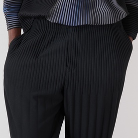 Model wearing Issey Miyake Basic Keshi Pleated Trousers in black, tapered leg with elasticated waistband, styled with striped top.