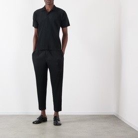 Model wearing Issey Miyake Pleated Wide Leg Trousers in black with elasticated waist, styled with a black polo shirt and black loafers.