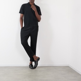 Model wearing Issey Miyake Pleated Wide Leg Trousers in black with a black polo shirt and dark slip-on shoes.