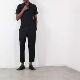 Model wearing Issey Miyake Open Collar Pleated Polo Shirt in black, styled with matching black tapered trousers and black loafers.