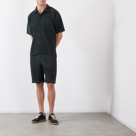 Model wearing Issey Miyake Special Black shorts styled with matching short-sleeve shirt and black low-top trainers.