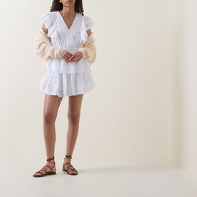 Model wearing LOVESHACKFANCY Gwen Heritage Mini Dress in white with tiered skirt and embroidered detail, styled with cream knit cardigan and brown