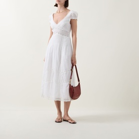 Model wearing LOVESHACKFANCY Suvi Lace Trim Flared Dress in white, midi length with V-neckline and cap sleeves, styled with brown bag and sandals.