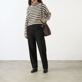 Model wearing Lisa Yang Zerlina Striped Turtleneck Sweater in Oyster/Dark Oak Stripes with black tailored trousers, carrying a burgundy bag.