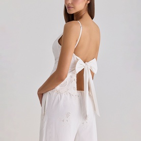 Model wearing House of CB white camisole top with open back, bow tie detail, and floral embroidery, styled with matching white wide-leg trousers.