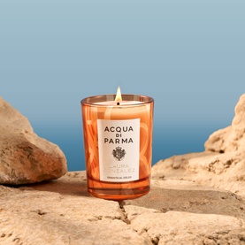 Acqua Di Parma beach candle in amber glass vessel with lit flame, styled on sun-warmed rocks against a blue sky backdrop.