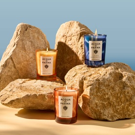 Acqua Di Parma candle trio displayed on sandy rocks, featuring amber, blue and orange glass vessels in a sun-drenched coastal setting