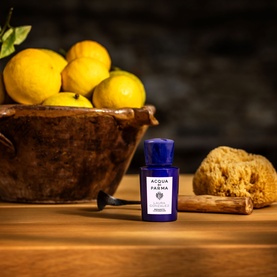 Acqua Di Parma blue EDP miniature bottle on wooden surface, styled with lemons in rustic bowl and natural sponge.