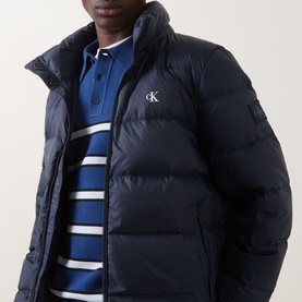 Model wearing Calvin Klein Jeans Dark Sapphire puffer jacket with high collar, styled with a blue striped polo shirt.