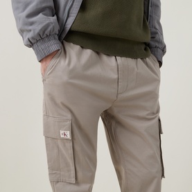 Model wearing Calvin Klein Jeans Comfort Skinny Leg Cargo Trousers in Vintage Khaki, with logo badge on side cargo pocket. Styled with green hoodie.