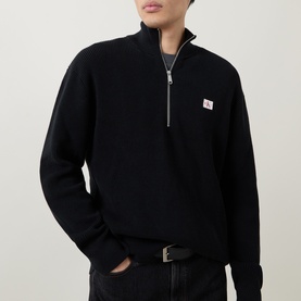 Model wearing Calvin Klein Jeans Badge Logo Waffle-Knit Half-Zip Sweatshirt in black, with funnel neck and chest badge logo, styled with a black belt.