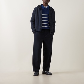 Model wearing navy zip-up jacket over blue striped polo shirt and black wide-leg trousers, styled with black leather loafers