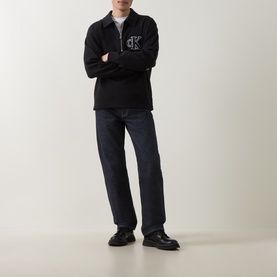 Model wearing black zip-up sweatshirt with chest logo, styled with dark indigo jeans and black chunky loafers
