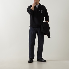 Model wearing black Calvin Klein half-zip sweatshirt with logo, styled with dark straight-leg jeans and black chunky loafers, carrying jacket