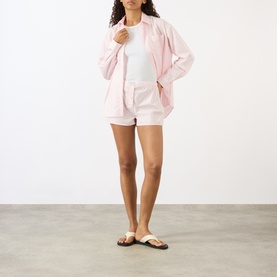 Model wearing Sporty and Rich Signature Logo Ring Shorts in Clear Pink with matching shirt jacket and white vest, styled with beige sandals.