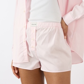 Model wearing Sporty and Rich Signature Logo Ring Shorts in Clear Pink with button fastenings and logo patch, styled with matching pink shirt.