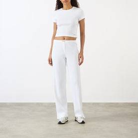 Model wearing Sporty and Rich Signature Logo Rib Baby T-Shirt in white, styled with matching wide-leg white trousers and grey trainers.